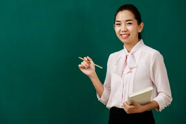 アジアの若い女性の美しい学校の先生の教師の先生の肖像画のクローズアップ笑顔立っている教授は教室の大学で黒板の前で手に鉛筆や教科書を保持カメラを見て — ストック写真