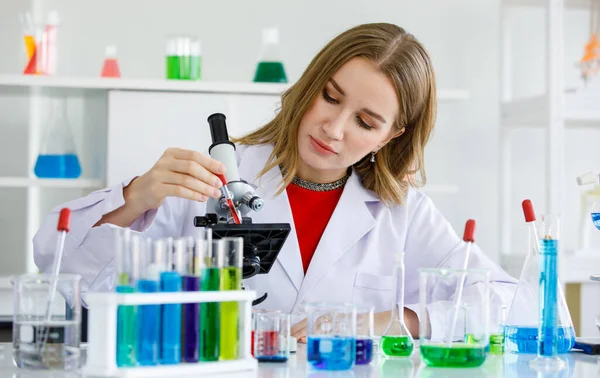 女性の白血病は科学者です前景と顕微鏡の近くに滴の液体を保持し 見て実験室で多くの試験管と化学試薬です — ストック写真