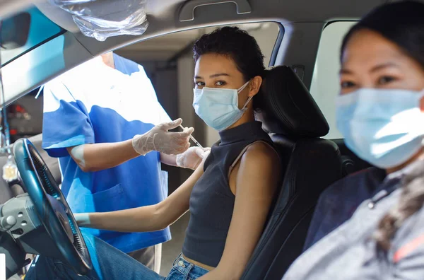 Female citizens wears face mask sitting look at camera in car with friend in drive through vaccinating line in hospital receiving coronavirus vaccine injection shot from public health doctor.
