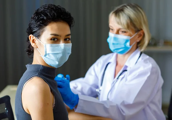 Beautiful short black hair female patient wears face mask sit look at camera while Caucasian doctor in white lab coat with stethoscope injecting vaccine in to her shoulder in blurred background.