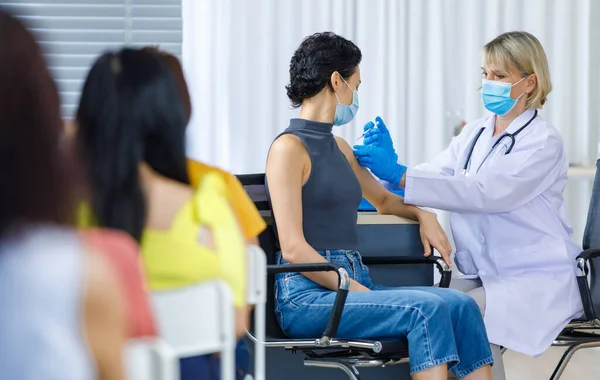 Cute female wears face mask sit look at camera while Caucasian woman doctor in white lab coat blue gloves and stethoscope injecting shot vaccine on shoulder when other patients wait in queue line.