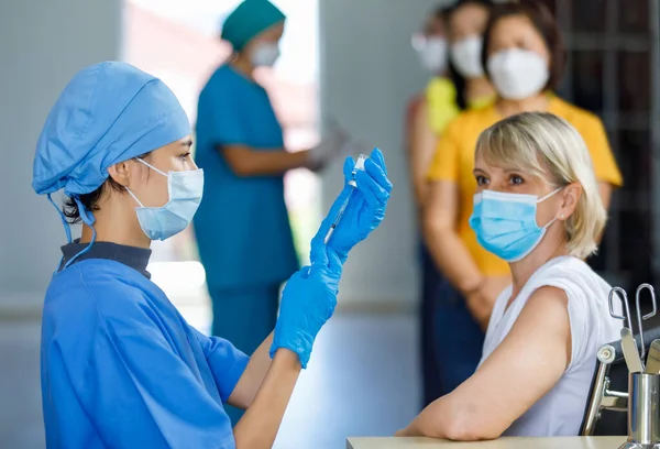 Cute doctor wears face mask rubber gloves and blue hospital uniform using needle syringe sucking coronavirus vaccine from small glass vial dose when Caucasian female patient wait for vaccination.