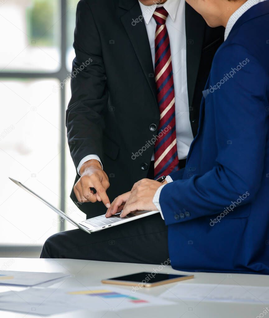 Cropped and unrecognizable two Asian male entrepreneurs standing and sitting in modern workplace with laptop and talking about new project