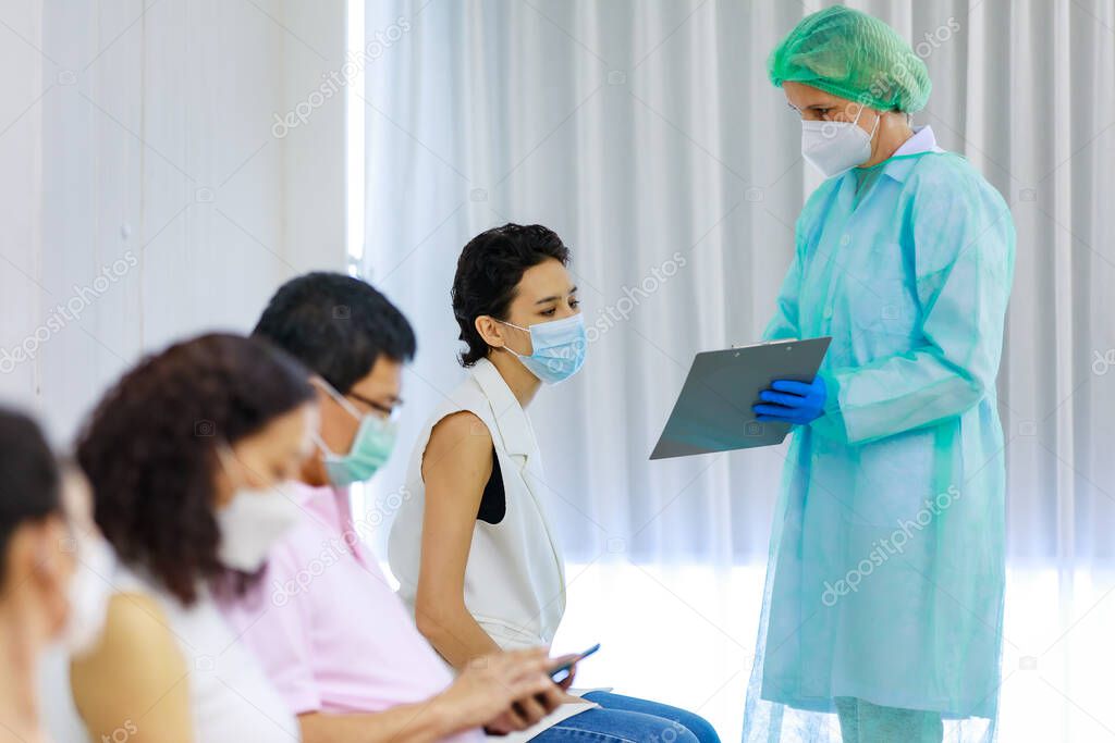 Young female citizen wears face mask in Coronavirus vaccinating queue line sitting checking her personal information from paper board in hand of full protective suit nurse who standing nearby.