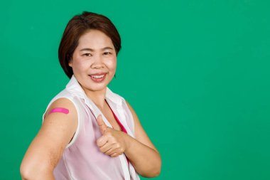 Orta yaşlı Asyalı kadın gülümsüyor ve başparmaklarını yukarı kaldırıyor. Kolunda bandaj bandıyla yeşil arka planda Covid 19 virüsü aşısı olduğunu gösteriyor. Covid 19 aşısı için kavram.