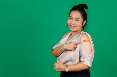 Asyalı orta yaşlı, gülümseyen ve koluna bandaj bandıyla bakan bir kadın yeşil arka planda Covid 19 virüsü aşısı olduğunu gösteriyor. Covid 19 aşısı için kavram.