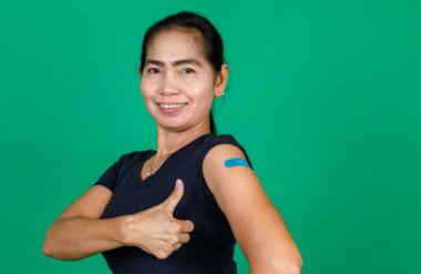 Asyalı orta yaşlı, gülümseyen ve koluna bandaj bandıyla bakan bir kadın yeşil arka planda Covid 19 virüsü aşısı olduğunu gösteriyor. Covid 19 aşısı için kavram.