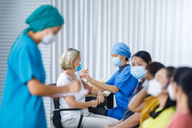 Kadın doktor yüz maskesi takıyor ve hastane üniforması giyiyor. Kafkasyalı yaşlı kadının omzuna koronavirüs aşısı enjekte ediyor. Diğer çok uluslu hastalar ise bulanık ön planda sırada bekliyorlar..