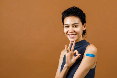 Coronavirus Covid-19 aşısı vurulduktan sonra omuzundaki mavi alçıda el işaretini gösteren gülümseyen Asyalı, neşeli ve seksi kadın hastanın yakın çekim portresi..