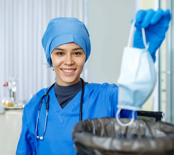 Closeup shot of female happy smiling doctor in blue hospital uniform rubber gloves and stethoscope look at camera dump used face mask into garbage bag trash can in blurred foreground after pandemic.