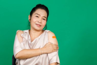 Kolunda bandaj bandı olan Asyalı orta yaşlı bir kadın yeşil arka planda Covid 19 virüsü aşısı olduğunu gösteriyor. Covid 19 aşısı için kavram.