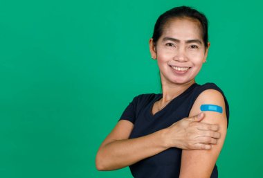 Orta yaşlı Asyalı kadın kolunu bandaj bandıyla göstererek yeşil arka planda Covid 19 virüsü aşısı olduğunu gösteriyor. Covid 19 aşısı için kavram.