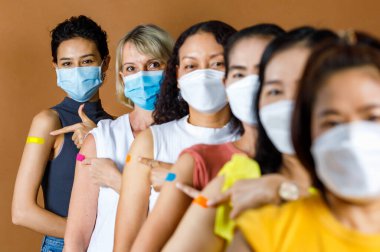Çok uluslu etnik kadın hastaların çeşitli grupları yüz maskesi takıyor boy sırasına göre sırada bekliyorlar. Aşılandıktan sonra omuzlarındaki renkli alçılara bakın..