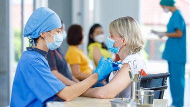 Asyalı kadın doktor yüz maskesi takıyor ve mavi hastane üniforması giyip koğuştaki yaşlı kadına aşıyı enjekte ederken diğer hastalar da bulanık arka planda sırada bekliyorlar..