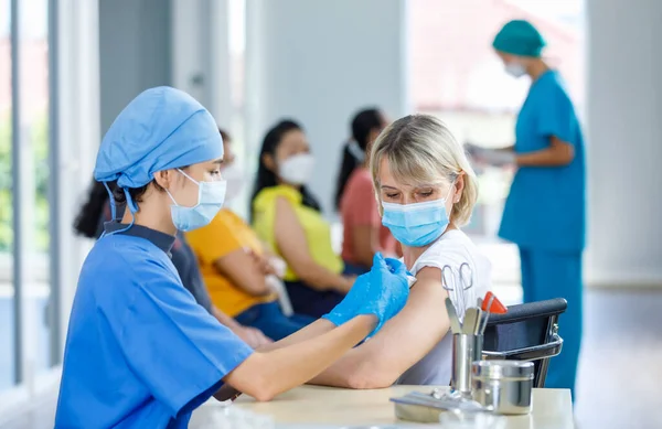 Asian female doctor wears face mask and blue hospital uniform injecting vaccine to Caucasian senior woman at ward working desk with equipment while other patients wait in queue in blurred background.