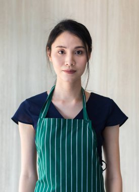 Portre, genç Asyalı güzel bayan baristanın, siyah gömlekli yeşil çizgili önlük giyip, kahverengi duvar kağıdı arka planındaki kameraya bakın..