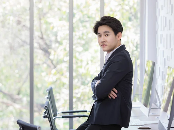 アジアの若いですハンサムな自信を持って成功したプロの男性実業家は 会社のカメラでオフィスの机の上に座って傾いて黒い正式なスーツを身に着けている — ストック写真