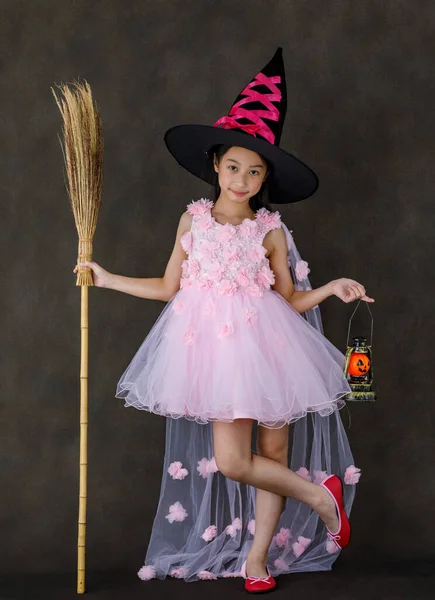 ピンクの魔女の衣装を着たかわいいアジアの子供の肖像画スタジオのショット 黒の高い帽子スタンドの笑顔が刻まれたオレンジ色のカボチャの顔のランプと魔法のブロムスティックハロウィンの伝統的な祭り — ストック写真