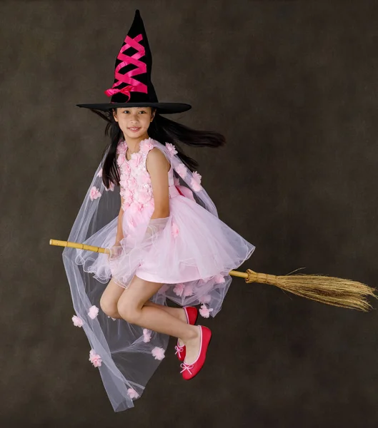 ポートレートスタジオショットの小さなかわいいアジアの子供でピンクの魔女の衣装で黒の高い帽子の表情でカメラポーズ飛行ジェスチャー乗って魔法の魔女ブロムスティック上のハロウィーンの伝統的な祭り — ストック写真