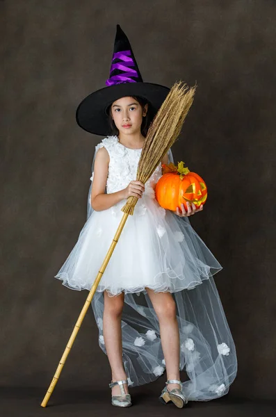 白い魔女の衣装で小さなかわいいアジアの子供のポートレートスタジオショット黒紫色の高い帽子立っている彫刻オレンジカボチャの顔と暗い背景の前に魔法のブロムスティック — ストック写真