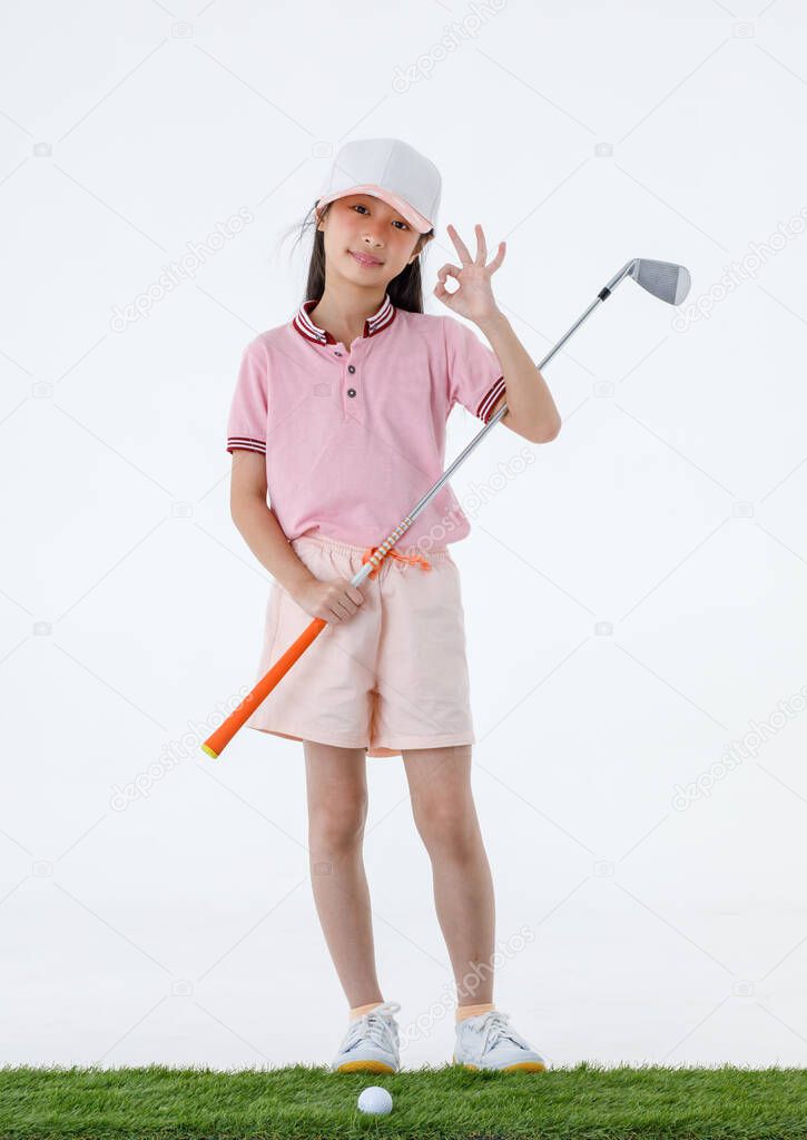 Studio shot of little Asian golfer in sport athlete uniform stand on green grass look at camera hold golf club driver in arm showing okay fingers sign in front of white background.