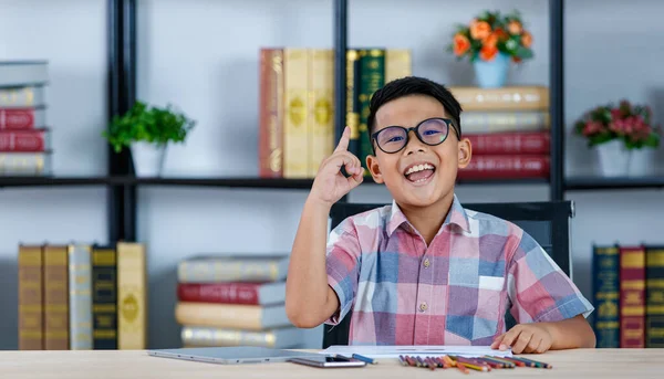 Smart young Asian boy wearing glasses and pink checked shirt sitting on desk of tablet, smart phone, and pencils in reading room and exciting on pop-up creative idea to complete homework of drawing