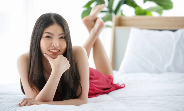 Beautiful young naughty sexy girl with long hair and red romantic underwear lying down on pillow at bed end seductively glance at someone with happy smile.