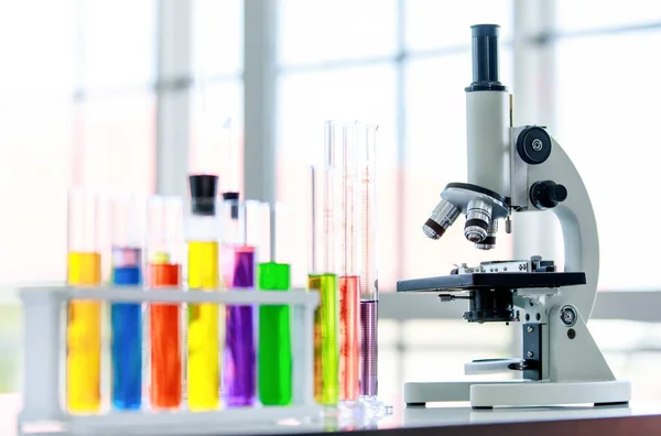 研究室のテーブルの上で3つのレンズ顕微鏡科学機器のクローズアップショットでは 色とりどりの試薬と溶液サンプルがぼやけた前景に卒業シリンダーを持つプラスチックラック内の試験管で — ストック写真