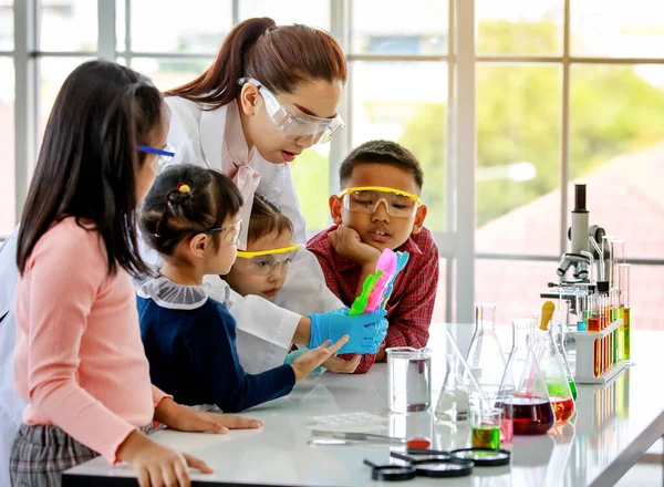 亚洲小学科学女教师身穿实验室外套 手里拿着五颜六色的实验室设备 教小男孩和小女孩戴着安全护目镜 如何在化学实验中使用它们 — 图库照片