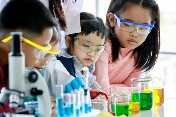 亚洲小女孩好奇地看着老师从玻璃杯中倒入红色试剂 用显微镜和设备与年轻朋友一起在实验室桌上做化学实验 — 图库照片