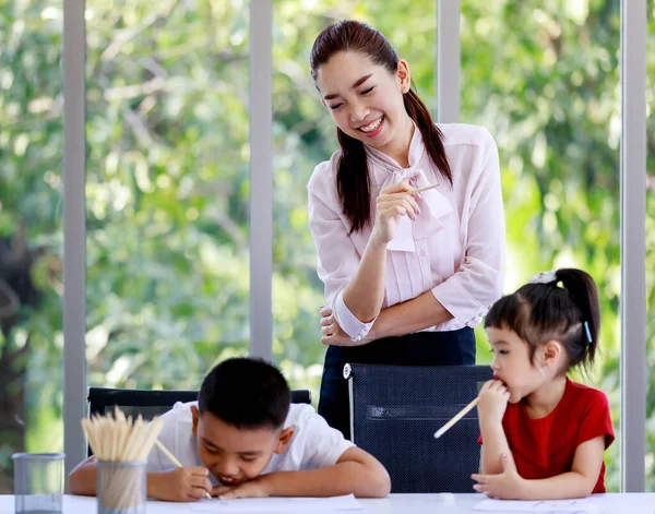ポートレートクローズアップアジアの幸せな美しい女子小学校教師笑顔立っているとき 小学校の男の子と小さな就学前の女の子を教える一緒にテーブルの上で宿題をする — ストック写真