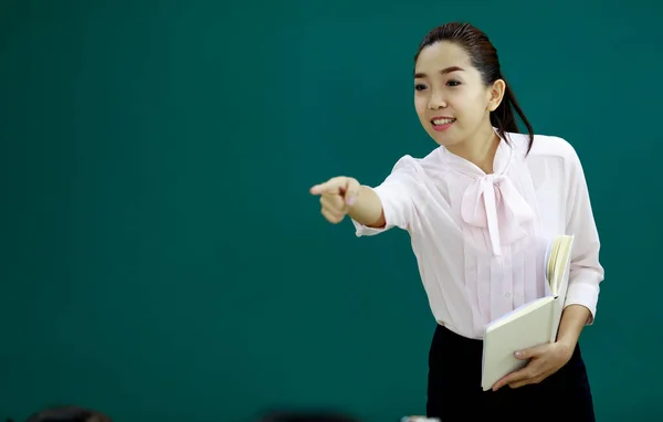 Portrait shot of Asian beautiful ponytail female teacher lecturer tutor standing smiling holding text book in hands pointing at children in front of green chalkboard in primary school lecture room.