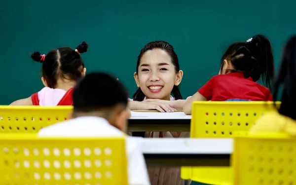 Asian young beautiful tutor teacher smiling sitting arming on table face to face in front of little cute preschool girl teaching helping her do exercise in classroom with other classmates in school.