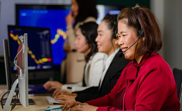 Group of Asian female customer service operators wears microphone headsets sit smiling in front daily graph chart computer monitor screen talking and trading buy sell stock market online with client.