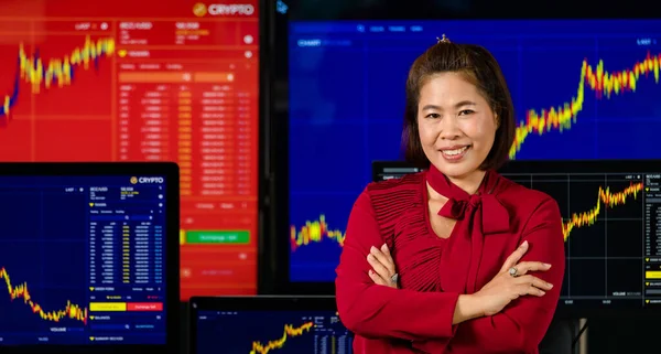 Asian female successful professional broker trader investor stand smile crossed arm look at camera in front computer screen with stock and bitcoin cryptocurrency graph chart analysis.