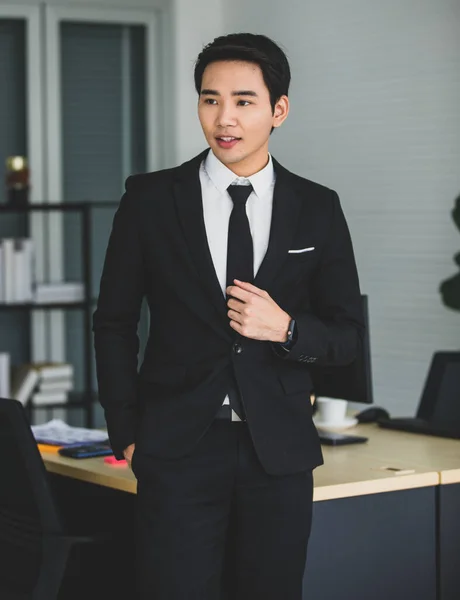 Modern businessman formal wear in black. Standing smiling right hand in pant pocket looking beside in office room. Looks friendly confident leadership relaxed style. Concept Young owner, intelligent