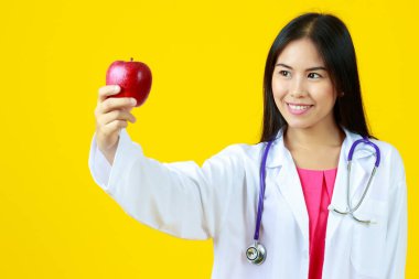 Asyalı genç, güzel bayan doktor steteskoplu beyaz önlük giyiyor, gülümsüyor ve kırmızı elmaya bakıyor. Beslenme, vitamin, sağlıklı ve izole edilmiş sarı arka planı sunar.
