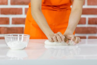 Turuncu önlüklü tanınmayan kadın gülümseyecek ve hamur yoğurmaktan zevk alacak. Cam kase un tozunun yanında beyaz yemek masasında yemek hazırlarken ev mutfağındaki tuğla duvarın yanında.