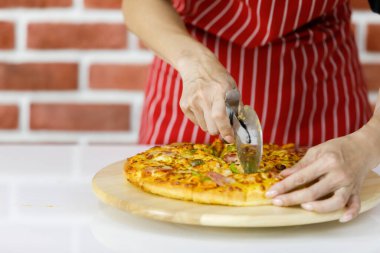 Kırmızı çizgili önlüklü kadın mutfaktaki ahşap tabakta geleneksel İtalyan yemeklerini yemek için lezzetli fırınlanmış pizzayı dikkatlice dilimlemek için jilet kullanıyor.