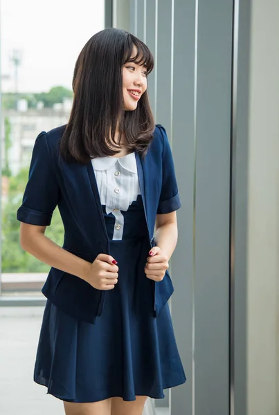 窓の前に立ち オフィスで休憩時間に外の木を見て暗い青の服でスタイリッシュな髪型をしたかわいい笑顔の若いアジアの女性の垂直肖像画 — ストック写真