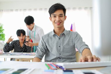 Asyalı erkek zanaatkar atölyede terzilerin yanında otururken masaüstü bilgisayarıyla çalışıyor ve kameraya bakıyor..