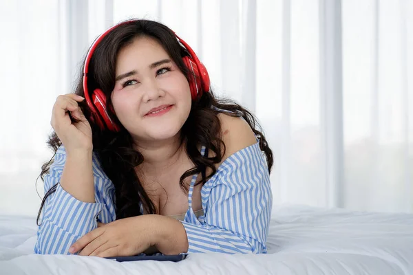 Asian young happy calm fat overweight woman lay down with happy smiling deep emotion peacefully while listening to music from big red earphones use playlist from black tablet at home in bedroom.