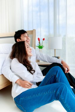 Genç ve çekici bir Asyalı çift yatak odasında birlikte oturup beyaz perdeden mavi gökyüzü manzarasına bakıyor. Aşk ve mutlu ilişki kavramı.