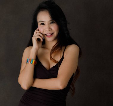 Uzun saçlı, mavi tırnaklı, siyah elbiseli, renkli aksesuarlı, koyu gri arka planda gülüp kameraya bakan hoş bir Asyalı kadın.