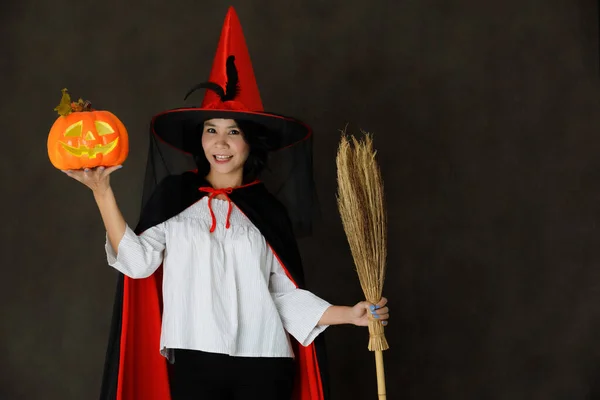 魅力的な笑顔若いですアジアの女性で魔女衣装でハロウィンカボチャとほうき見てカメラで暗い灰色の背景 — ストック写真