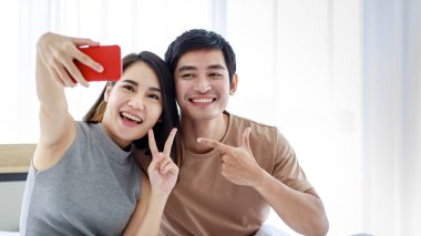 Neşeli genç Asyalı kadın ve erkek, evde birlikte vakit geçirirken cep telefonuyla selfie çekiyorlar.