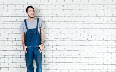 Önlüklü genç adam, elinde fotokopi aletiyle beyaz tuğla duvarın önüne bakıp gülümsüyor..