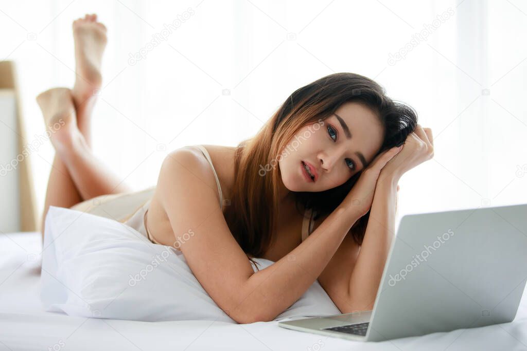Young sexy Asian woman wearing underwear leaning on hand and browsing laptop while relaxing on bed in light bedroom in morning. Private time ralax concept.