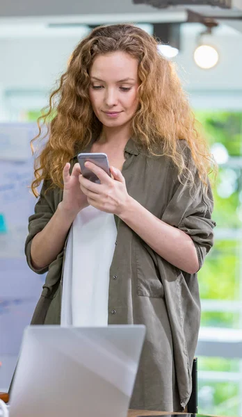 Caucasian creative businesswoman wearing casual hipster clothes for working, using and holding mobile phone, touching screen for chatting and gently smiling while standing in indoor modern office