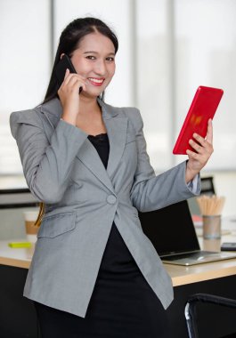 Gri takım elbiseli genç çekici Asyalı kadın cep telefonuyla konuşurken modern görünümlü ofiste bulanık pencereli tablete bakıyor. Modern ofis yaşam tarzı kavramı.